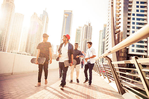 Friends enjoying Dubai city life Group of happy friend spending some time together in Dubai Marina during their summer vacation. The sunlight is on the background and having fun embracing and smiling walking in the streets. Two guys on front holding a skateboard, the guy on the right wearing a red hat. dubai street stock pictures, royalty-free photos & images