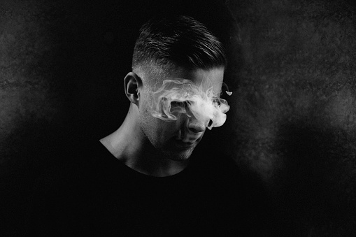 Black and white portrait of young man, smoke hiding eyes. Film grain added.