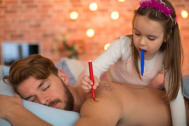 Cute daughter drawing on her sleeping father's back. Cute daughter drawing butterfly and rainbow with felt pen on her sleeping father's back. Father is lying on stomach with eyes closed, while girl wearing princess costume. happy fathers day funny stock pictures, royalty-free photos & images