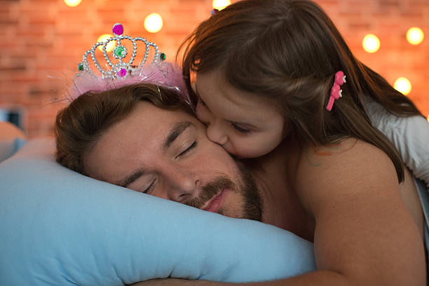 Cute daughter kissing her sleeping father. Cute daughter kissing her sleeping father. Father is lying on stomach in bed with eyes closed, wearing princess tiara. happy fathers day funny stock pictures, royalty-free photos & images