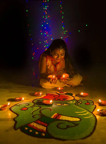 A Little girl making Rangoli and decorating with Oil lamps for Diwali celebration in India. The rangoli design is that of a traditional elephant head depicting Lord Ganesha. The girl is holding diyas(oil lamps) in her hands. Diwali lights are seen in the background.