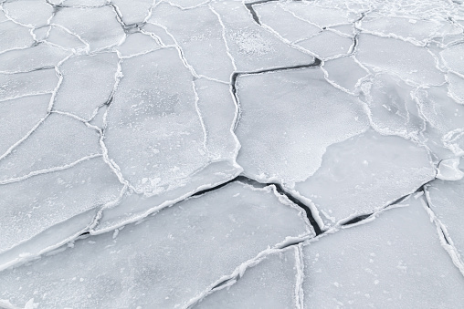 Detail of cracked, frozen sea ice in Ilulissat, Greenland. The image is predominantly white, with dark grey showing through the edges of the ice sheets. Glimpses of the sea can be seen through the zig-zag pattern of the ice. On this day with the wind chill, it was about -42 degrees C.