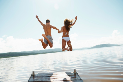An excited young couple holding hands while jumping off the jetty at the lake