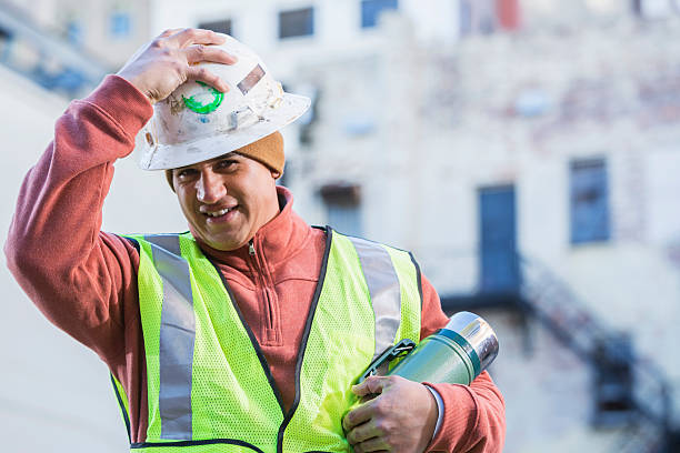 A mid adult, Hispanic man putting an old white hard hat on his head.  He is wearing a yellow safety vest and holding a thermos, standing and smiling at the camera.  He is a repairman or construction or utility worker, working in the city streets with buildings out of focus behind him.