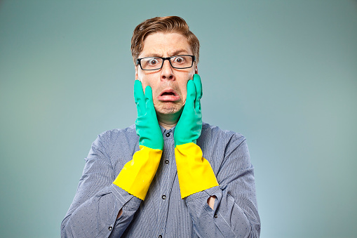 Nerdy guy with green and yellow rubber gloves on looking shocked, with his hands on his face looking at camera. Something needs cleaning. 