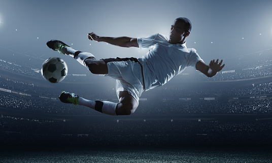 A male soccer player makes a dramatic play by jumping horizontally. He attempts to kick the ball with his feet. The stadium is dark behind him. Only the lights of the stadium shine brightly, creating a halo effect around the bulbs.