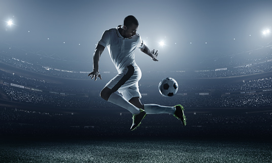 A male soccer player makes a dramatic play by jumping horizontally. He attempts to kick the ball with his feet. The stadium is dark behind him. Only the lights of the stadium shine brightly, creating a halo effect around the bulbs.