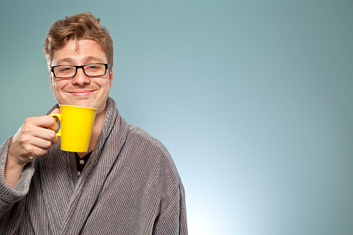 Nerdy guy sipping coffee and looking blissful in the early hours of the morning. He's wearing a dressing gown and is still in his pyjamas having just gotten out of bed.
