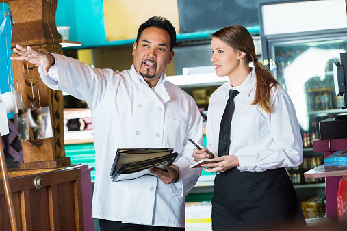 Mature Hispanic man wearing chef's coat is holding menus and gesturing into restaurant dining room. He is training a new female waitress who is wearing a traditional uniform and apron. They are standing in front of a commercial kitchen in a Tex-Mex or Mexican restaurant. Chef is holding menus. 