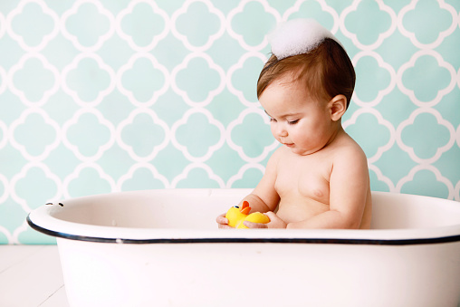 A baby looks at a rubber ducky as she sits in an antique bathtub. Copy space in vintage wallpaper. 