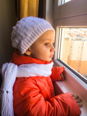 close up shot of little girl looking out from window with coat and scarf waiting her father