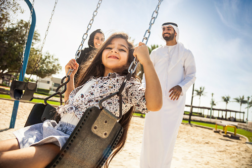 Happy young traditional family in Dubai, UAE at the park. The little girl playing on the swing with her dad and mom.