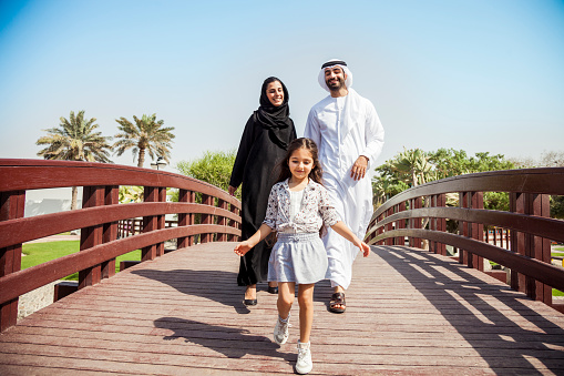 Happy young traditional family in Dubai, UAE at the park. The little girl running in front of their parents in a beautiful sunny day.