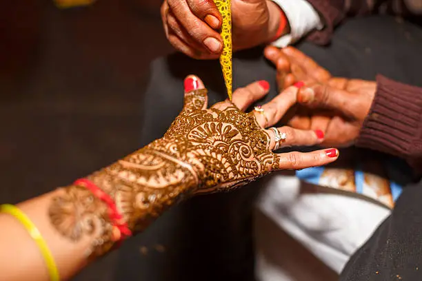 Henna Tattoo or mehendi pattern being applied on an Indian bride's hand. The bride has henna paste designs or patterns on her hands as decoration and a beauty feature along with the other adornments. The image shows the forearm of the bride, being held by the tattoo maker and Mehendi being applied from a plastic cone.
