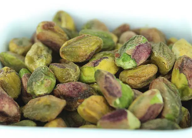 Photo showing a small white dish that has been filled with a pile of green, pistachio seeds / nuts.  The kernels have all been shelled and are ready to be eaten raw, while some prefer them to be roasted or salted.  Pistachios not only taste delicious, but they also boasts many health benefits, helping to improve your circulation.