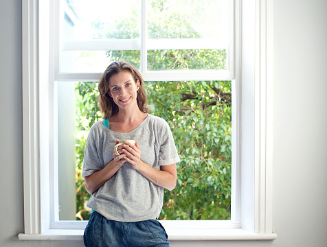 Portrait of a relaxed mid adult woman smiling with cup of coffee by window at home