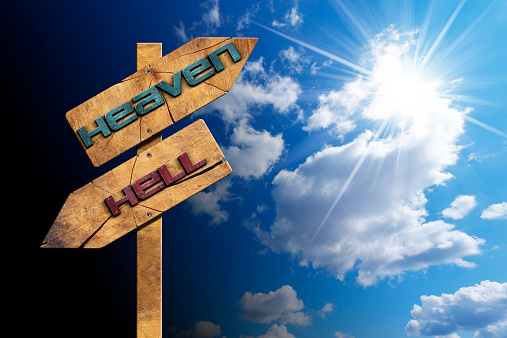 Wooden directional sign with two arrows in opposite direction with text heaven and hell on blue sky with clouds and sun rays