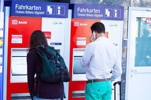 Münster, Germany - May 31, 2014: Two young caucasian adult people, a woman and a man, are standing at two ticket machines of german Deutsche Bahn in hall of station Münster. They are buying tickets. Man is using mobile at same time.