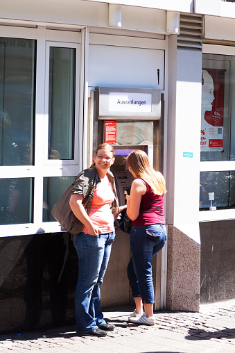 Münster, Germany - May 31, 2014: Capture of two young caucsian women at ATM in sunshine in Münster. One girl is using ATM. Other girl is watching into street.