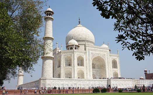 Agra, India - March 22, 2015: The Taj Mahal, crowded with tourists and local visitors. The building is a white marble mausoleum located in Agra, Uttar Pradesh, India. It was commissioned in 1632 by the Mughal emperor Shah Jahan to house the tomb of his third wife, Mumtaz Mahal and stands on the south bank of the Yamuna River. It is possibly the best example of Mughal architecture in the world, blending elements of Islamic, Persian, Ottoman Turkish and Indian architectural styles. It is a major Indian tourist attraction attracting more than two million visitors a year. Foreground foliage. Unidentified people.