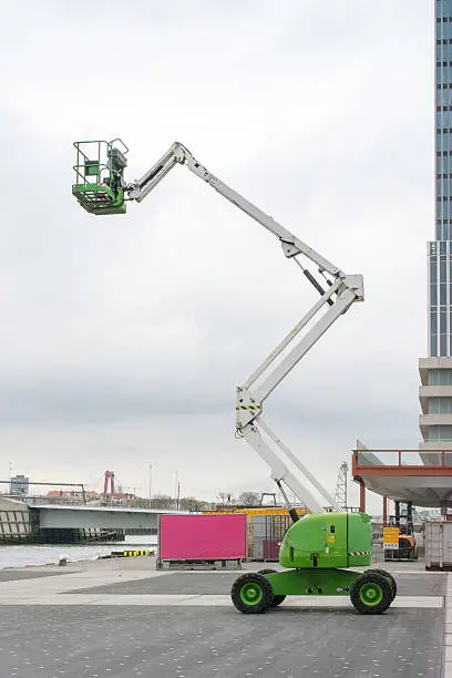 full length upright white and green empty cherry picker in front of large office tower