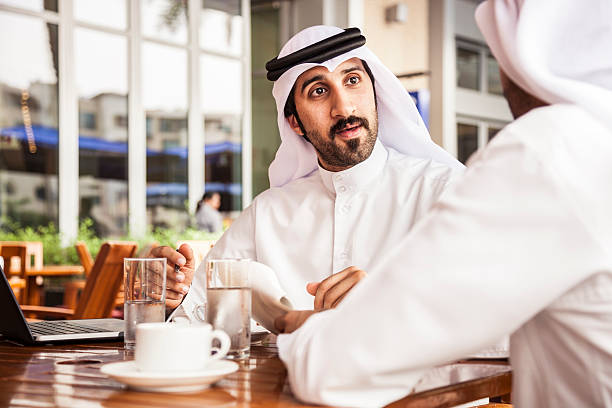 Two emirates business people wearing traditional dish dasha clothing doing business in financial district in Dubai, UAE. They are sitting in a cafe filling documents.