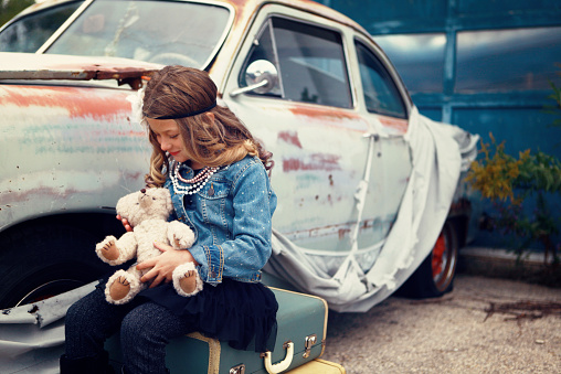 A little girl, seven years old, plays with her teddy bear as she sits on vintage suitcases beside an antique car. Shot with a Canon 5D Mark II. 