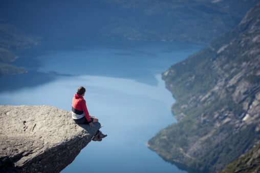 man sitting on trolltunga rock above lake in norway