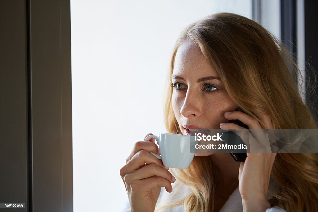 Young woman talking on the phone and drinking coffee Young woman talking on the phone and drinking coffee at the window 2015 Stock Photo