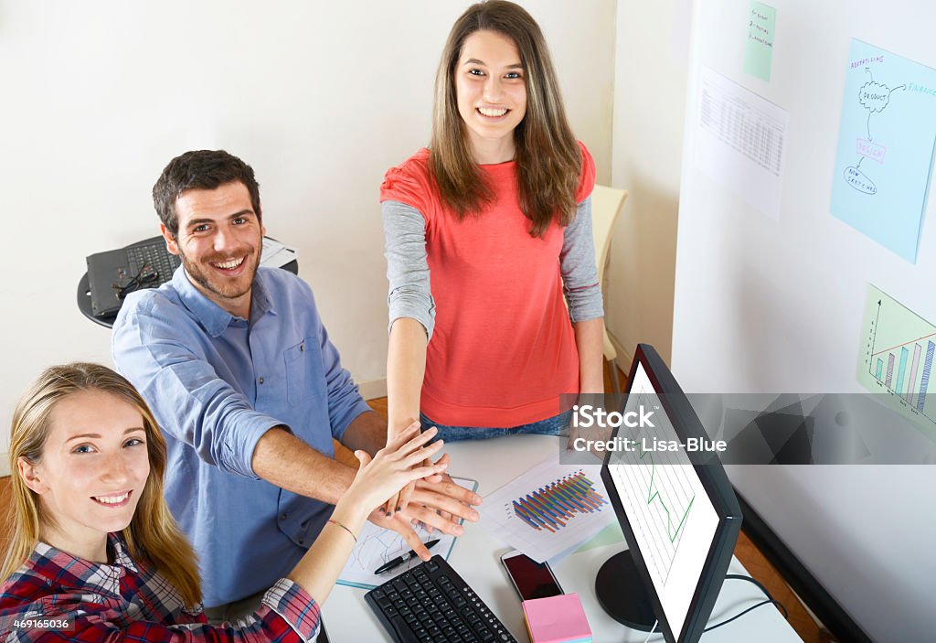 Teamwork Businessman or Design Professional and Colleagues stacking hands upon the desk in office. They have papers,pc,smartphone and reports. On computer monitor there is a chart. In the background there is a whiteboard with adhesive notes,charts and reports. They are looking at camera. High angle view. 2015 Stock Photo