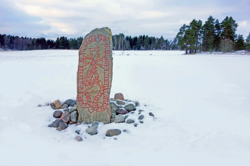 A picture of a swedish runestone standing in a winter landscape field, showing woods in the background.