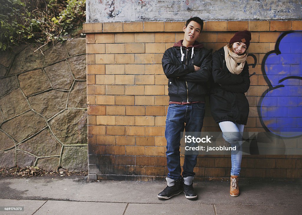Attractive teenage couple together outdoors Beautiful young man and woman in warm clothes standing together leaning on a wall outdoors. Teenage asian couple posing at camera on street. Adolescence Stock Photo