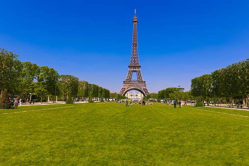 A digital photograph of the Eiffel Tower structure in Paris, France, as viewed from the Champ-de-Mars in perspective with clear blue, cloudless, skies serving as background during a vibrant sunny afternoon. Few people in the photo allowing for a clearer view of the grass and fading treeline that give in to the tower.