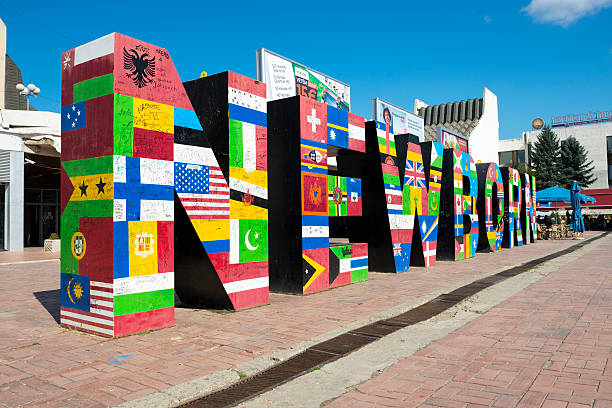 Pristina, Kosovo - August 29, 2013: The NEWBORN monument, which is located in front of a shopping center in Pristina, was unveiled when Kosovo declared independence on 17 February 2008. The flags of nations that recognized the country's independence are displayed on the monument. People at a cafe are visible at the end of the monument.