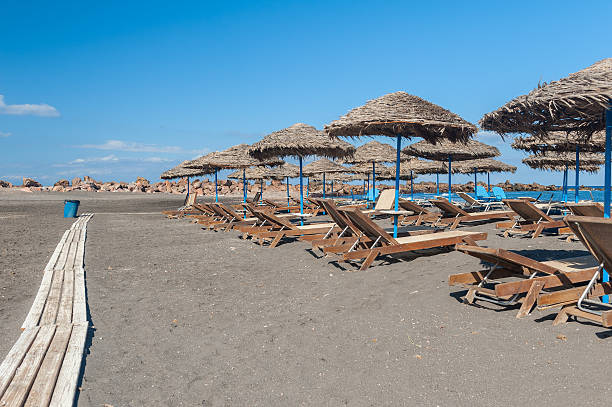 Gray sand beach with chairs, parasols and wooden walkway. Monolithos, Santorini, Greece.