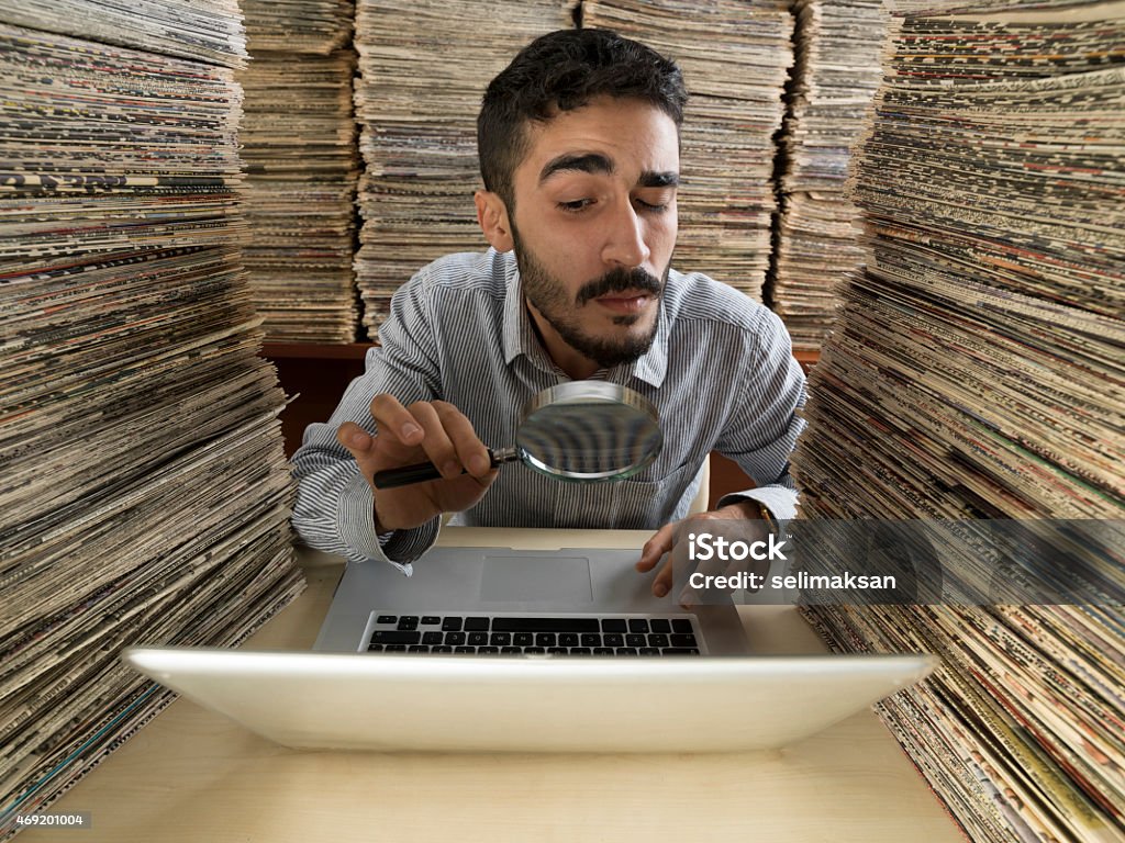 Adult man with dark hair doing research in media archive Adult man with dark hair looking through a magnifying glass for research in media archive full of large stack of newspapers.A laptop computer is on the desk.The model is wearing gray shirt, holding magnifying glass in right hand.Huge amount of real newspapers were used on the desk and on the background.The image was shot with Hasselblad H4D in horizontal composition. Magnifying Glass Stock Photo