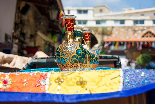 Old Phoenician vintage bottles made of stained colorful glass, embossed in gold, sitting a top of a beautifully-stitched tablecloth. De-focused in the background are the market buildings. The three vases are lined symmetrically, one behind the other, on the merchant table.