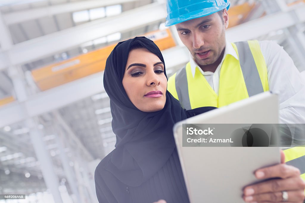 Checking Progres Together Woman architect wearing abaya, traditional Emirati clothes, on a construction site talking to a construction foreman. She is holding digital tablet. They are both looking at it. Construction Worker Stock Photo