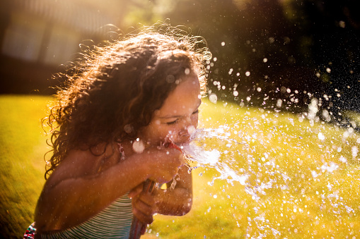 Little mixed race girl happily drinking water straight from a garden hose in her yard in summertime