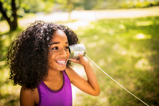 Young Afro girl listening happily to a tin can phone in sunny park