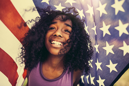 Proud and patriotic Afro girl smiling happily under an American flag, with cross process develop