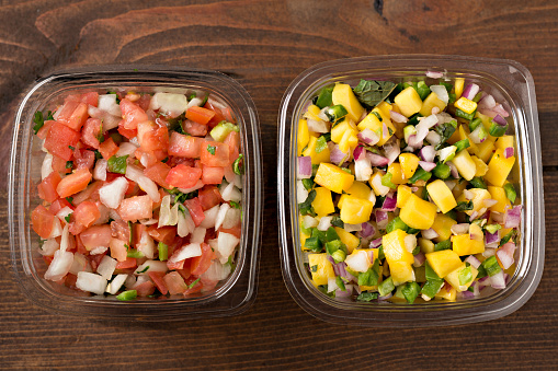 An overhead, close up horizontal photograph of two store bought plastic containers of spicy pico de gallo and mango salsa, two of the many prepackaged, labor saving foods available in your local grocery store.
