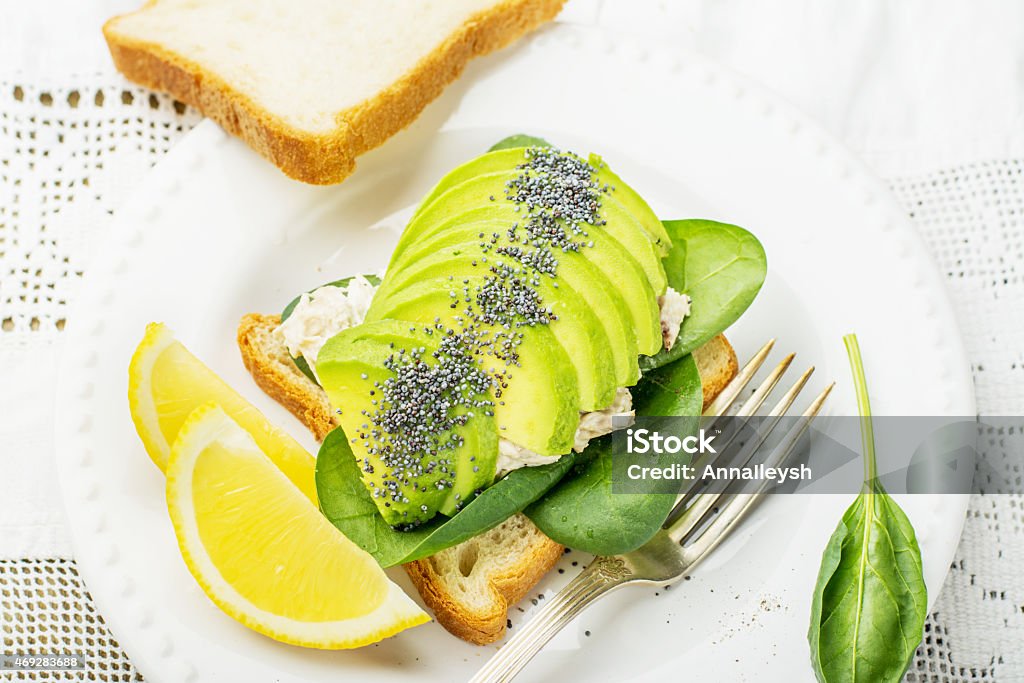 Sandwich  of slices  fresh white bread toast with spinach, chicken Sandwich of the pair of slices of fresh white bread toast with spinach, chicken, dressing, sliced avocado and poppy seeds. On a white plate and white vintage embroidered tablecloth. selective Focus 2015 Stock Photo
