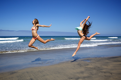 Two teenage girls in bikinis leaping in the air at the beach.  They are fit and athletic.  One is performing a firebird leap. Photo taken in San Diego in Southern California
