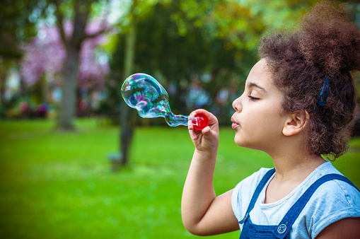 Stock photo of a mixed race girl who is blowing soap bubbles in playground. She is 8 years old, wearing blue overalls with a white t-shirt underneath. She is playing with a red bubble wand. In post-production, I increased contrast and vibrancy, added a vignette. This file has a 