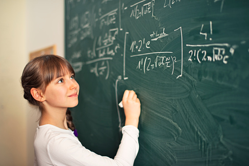 Little mathematics genius girl writing difficult mathematics equations on a green chalkboard. The gilr is smiling and obvioulsy she is having fun solving the equation.