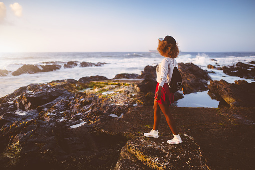 Afro teenage hipster girl standing looking out at the ocean on some rocks at the beach