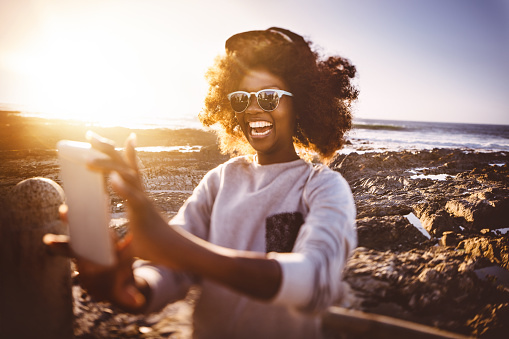 Happy African American hipster teen girl at the beach taking a selfie on her phone