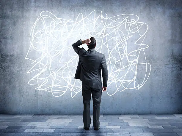 A confused businessman stares at a scribble design that is painted on a wall.  It represents the confusion and stress that some people feel in everyday life. He is looking up while standing with his back to the camera.