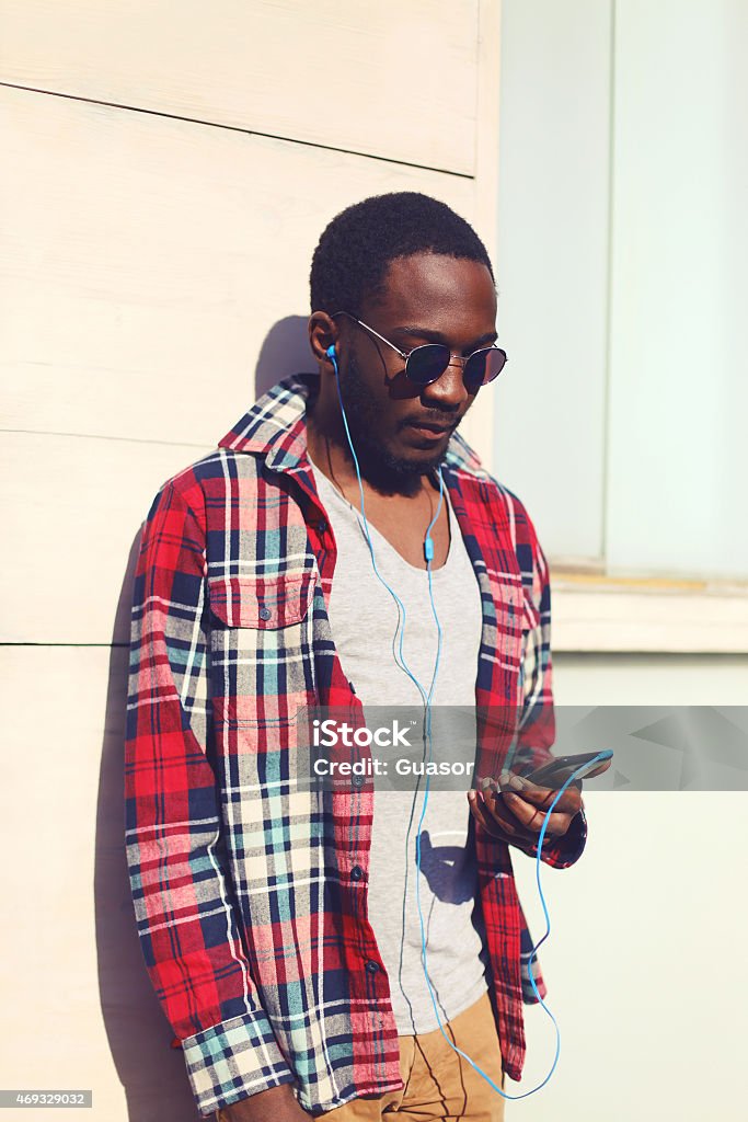 Fashion portrait of stylish young african man listens to music Fashion portrait of stylish young african man listens to music on the smartphone, hipster wearing a plaid red shirt and sunglasses against the urban wall outdoors 2015 Stock Photo